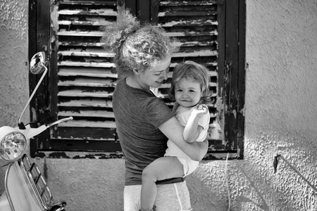 年轻美丽的金发女子捧着的小男孩婴儿在手站附近房子，有棕色的窗口和白色摩托车城市交通晴天室外 卧式图片
