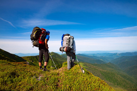 冒险家与背包站立在绿色山山脊反对蓝色天空的背景和敬佩山的全景在他们面前。史诗般的旅行在山上。后退视图
