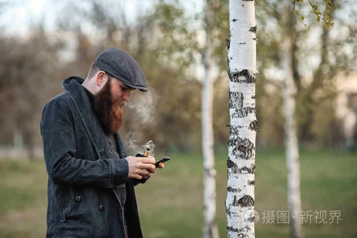 室外肖像的残酷白人小伙子留着大胡子和复古帽 vaping 一种电子香烟