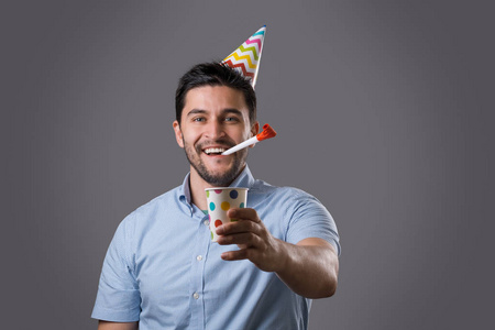 英俊的微笑年轻的深色家伙与党盖帽制造噪声由红色垫铁并且拿着纸杯子。新年或生日庆祝