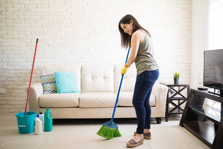 快乐的年轻女佣扫地板与绿色和蓝色扫帚在家里
