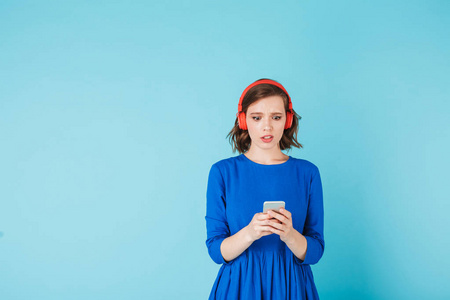照片的年轻 shoked 女士穿着和红色的耳机站在和惊异看着她的手机上的蓝色背景