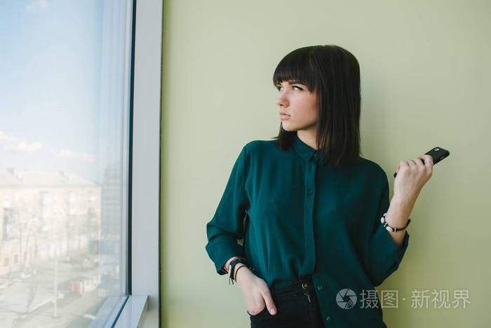 女青年商人,在一件深色的衬衫,手里拿站在现代办公室电话和寻找到窗口