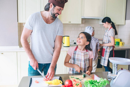 快乐的爸爸站在他的女儿和削减蔬菜。他们看着对方, 微笑着。妈妈 workswith 的儿子在炉子后面
