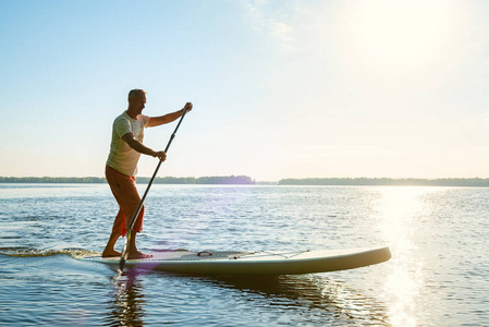 快乐的人是在阳光明媚的早晨, 在一条大河上的一个支撑板上训练。站起来桨登机真棒活跃的娱乐性质。背光, 侧面视图