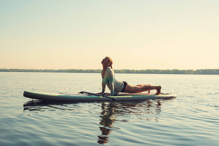 女性在一条大河上的阳光明媚的早晨练习瑜伽。站起来桨登机真棒活跃的娱乐性质。侧面视图