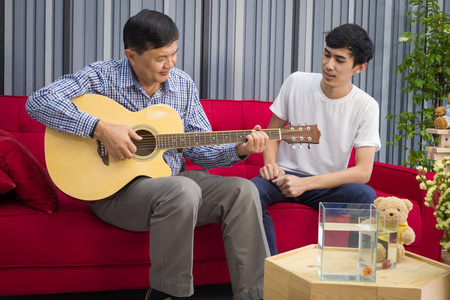 亚父子坐着弹吉他, 唱着建立家庭关系。快乐地在客厅在他们的家