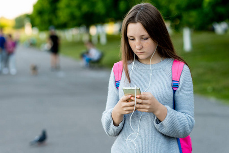 女孩少女在夏天在公园户外。在她的手中持有一款智能手机, 在耳机上听音乐, 在电话上写信息。文本的可用空间