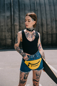 年轻迷人的纹身女孩手在口袋里拿着滑板在街上
