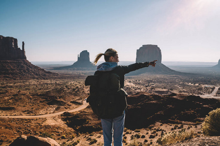 后视图年轻女性旅行者背包指向美丽的风景纪念碑谷与砂岩形成, 时髦女孩与背包徒步旅行在纳瓦霍保留探索独特的地质学