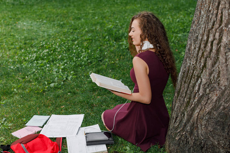 夏天公园里的卷发年轻漂亮女孩拿着书坐在树下