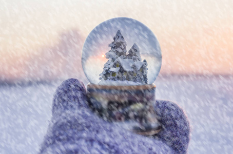 女孩在针织手套持有与 firtrees 房子和人造雪与冬季景观与背景下飘落的雪花玻璃球