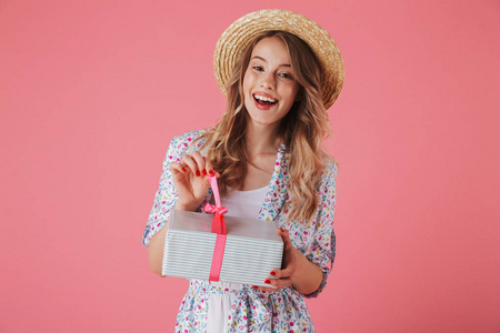 一个快乐的年轻女子在夏季礼服和草帽控股礼品盒在粉红色背景下隔离的肖像