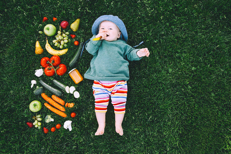 婴儿在五颜六色的衣服上尝试食物和框架的不同新鲜水果和蔬菜的绿色草背景, 顶部的看法。健康儿童营养, 婴儿喂养