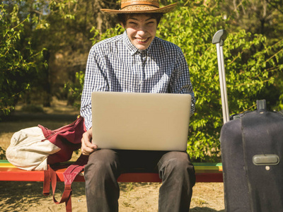 微笑的年轻人工作在笔记本电脑坐在长凳上与旅行箱和袋子