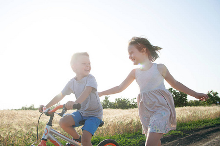 在田野里的男孩和女孩。男孩正在骑自行车, 女孩正在旁边跑。一个快乐, 幸福的童年在村里