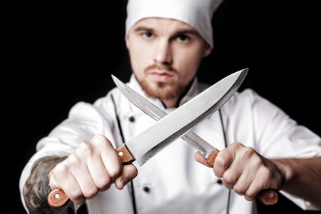 年轻胡子男人厨师在白色均匀搁置在黑色背景上的两把刀。专注于刀