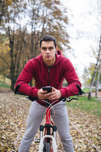 秋季户外活动。年轻严肃英俊的家伙与耳机站在公园的自行车, 手持手机和采摘音乐曲目