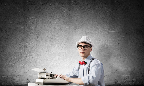 青年作家在帽子和眼镜使用打字机, 而坐在桌子上反对灰色混凝土墙的背景