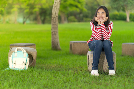 在阳光明媚的夏日, 在学校公园里坐着微笑的亚洲学生女孩