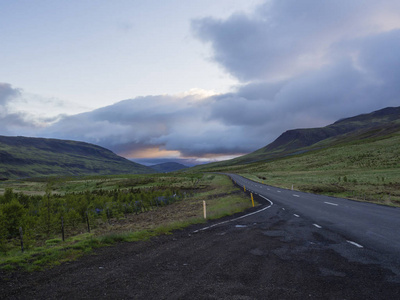 沥青道路曲线通过空北景观与绿草五颜六色的丘陵和日落戏剧性的天空, 方式到山在冰岛西部高地, 复制空间