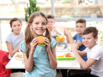 逗人喜爱的女孩吃汉堡和孩子坐在桌在学校自助餐厅