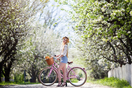 美丽的金发女孩拿着一篮子花的自行车, 微笑着甜美的微笑。这个女孩穿着白色的夏日生活。