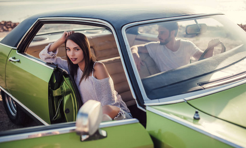 浪漫情侣坐在绿色复古车在海滩上。英俊的胡子男子和漂亮的年轻女子在老式的经典轿车。时尚爱情故事