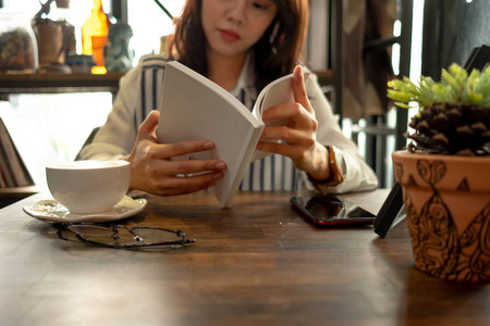 生活背景的年轻女子坐在餐厅和阅读书与杯咖啡, 眼镜和植物放置在木桌上