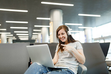 带耳机的旅行者妇女在笔记本电脑上用手指指着食指在机场大堂大厅等候。周末旅客出国旅行。航空飞行概念