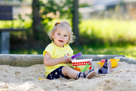 在户外操场上玩沙子的可爱的学步女孩。美丽的婴孩在红色胶长裤有乐趣在阳光温暖的夏天天
