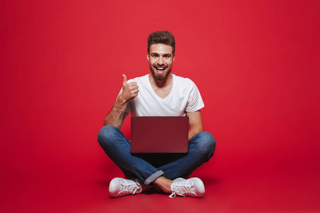 一个快乐的年轻胡子男子的肖像显示竖起大拇指的手势, 而坐在笔记本电脑孤立的红色背景