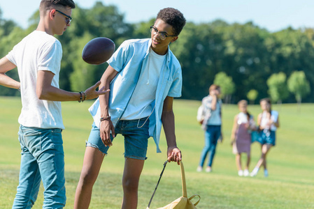 十几岁的多民族朋友玩橄榄球, 而同学们在公园后面走着