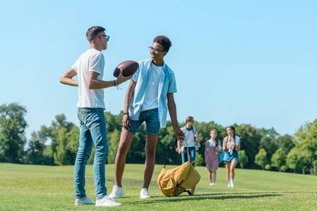 多民族男孩玩橄榄球, 而同学们在公园后面走