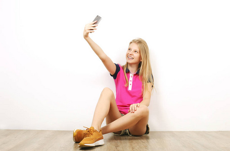 美丽的少女坐在木地板上拍摄自拍的手机。穿着黄色运动鞋的休闲年轻女性自己在智能手机上拍照, 靠在墙上。背景, 复制空间