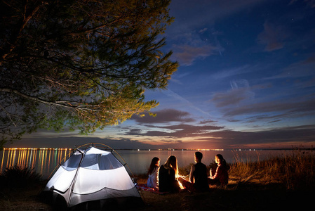 一群四游客坐在海边的篝火前的旅游帐篷树下, 胡子男子指着明亮的蓝色傍晚的天空, 在清澈的水背景。旅游业和野营概念