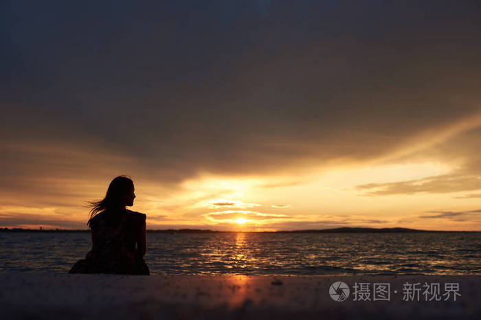 旅游妇女的背影剪影独自坐在海边的水边, 欣赏美丽的日落景色在黑暗的