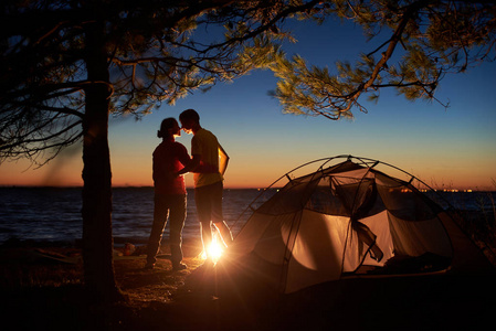 夜间露营在树下的海上。登山夫妇的剪影, 男子和妇女的后视图站在篝火旁, 晚上拥抱附近的旅游帐篷。旅游快乐关系与户外露营理念