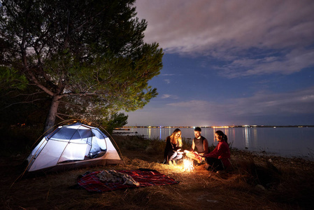 三名游客年轻人和妇女坐在湖岸边的篝火旁, 在树下的旅游帐篷附近。安静的水面和傍晚的天空背景。旅游, 友谊和露营概念
