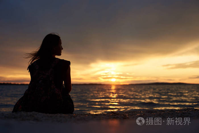 后景色旅游妇女的剪影与背包独自坐在海边的水边, 享受美丽的日落景色