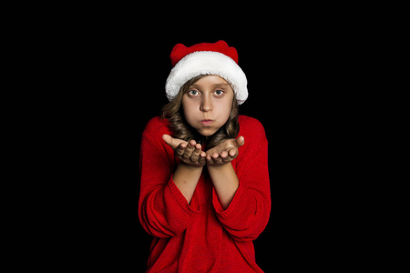 一个穿着红色毛衣和圣诞老人帽子的年轻女孩在她的手上吹着, 看着观众。黑隔离。复制空间