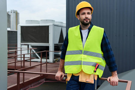 男建筑师在安全背心和安全帽的工具带站在屋顶上