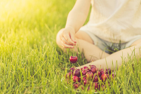 一个小女孩坐在绿色的草坪上, 从一盘甜樱桃上拿出成熟的浆果, 站在她的脚下。选择性聚焦
