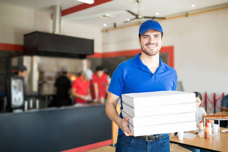 英俊的比萨送货男子信使在蓝色制服与帽子举行比萨饼盒微笑着看着相机