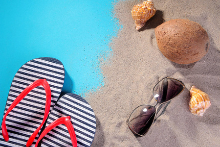 沙滩配件。拖鞋, 太阳的眼镜, 贝壳和椰子在海边的沙滩上。在蓝色背景下。顶部视图