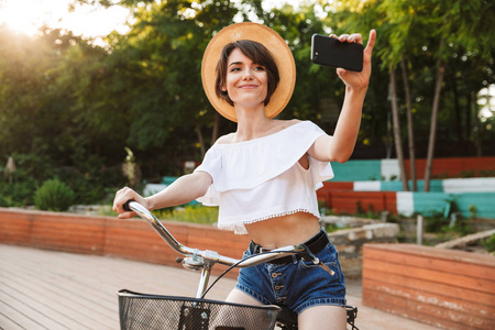 微笑的年轻女孩在夏天衣裳在公园骑自行车, 采取自拍与移动电话