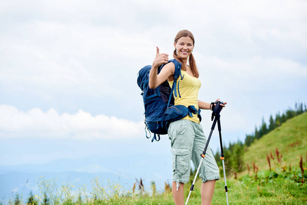 迷人的快乐女人背包徒步登山小径, 走在草地上的山上, 穿着背包, 使用徒步棍, 微笑着对相机和显示竖起大拇指。户外活动, 旅游理