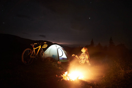 在夜间露营的年轻女子旅行者在燃烧的篝火旁野营, 照亮旅游帐篷, 在美丽的夜空下布满繁星的山地自行车。户外活动与旅游理念