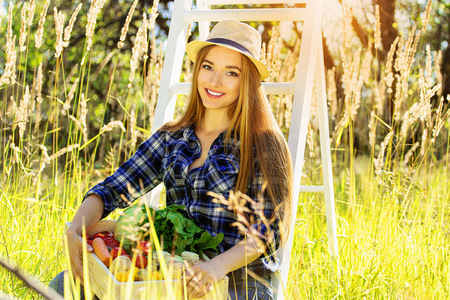 漂亮和年轻小女孩微笑着在帽子里保持装满蔬菜的木箱子。夏收