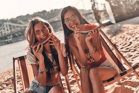 两位美丽的年轻女子坐在沙滩上微笑着吃西瓜
